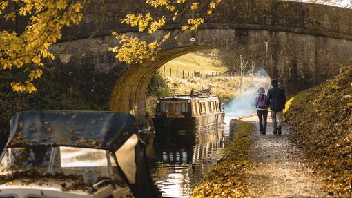 A couple walking beside the canal approaching a low bridge in Bathamption, Somerset, England