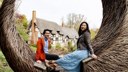 Asian couple sitting on a wooden sculpture in Stratford-upon-Avon with Shakespeare's birthplace in the backdrop