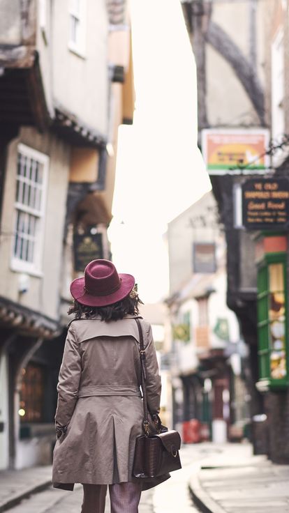 Woman wearing trench coat and pink hat walking through narrow historic street of York, North Yorkshire, England. 