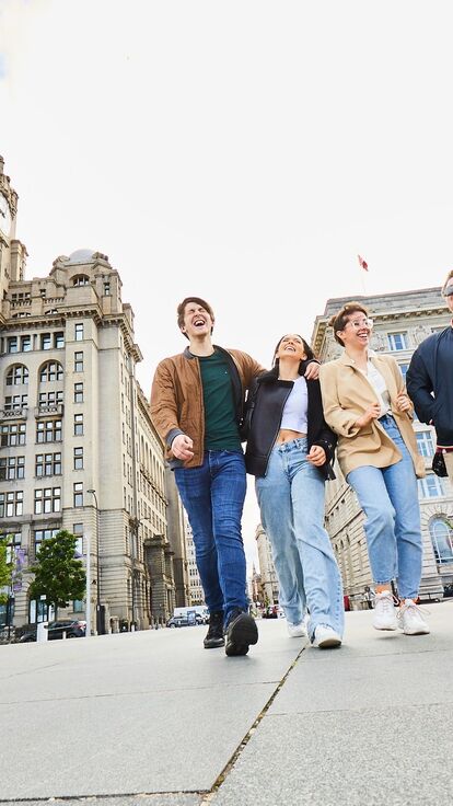Four friends exploring Liverpool walking along waterfront promenade, with historic buildings in the background