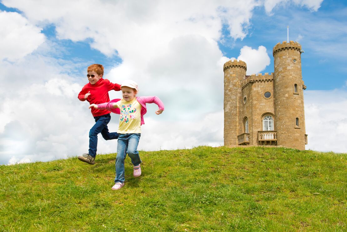 Two children running down a hill near a stone tower in the Cotswolds