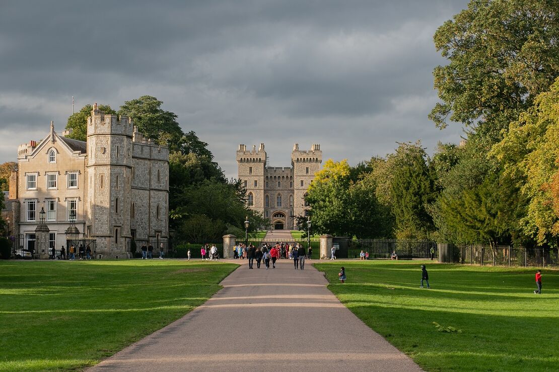 People walking down a path in front of a majestic castle, surrounded by lush greenery and a clear blue sky.
