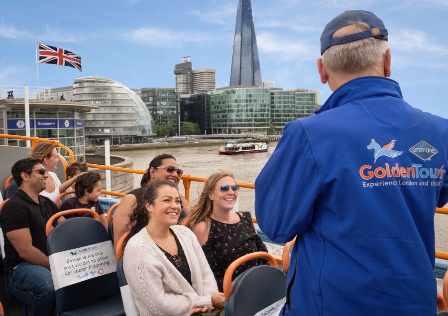 A tour guide engages in conversation with a group of people aboard the tour bus over the Thames River