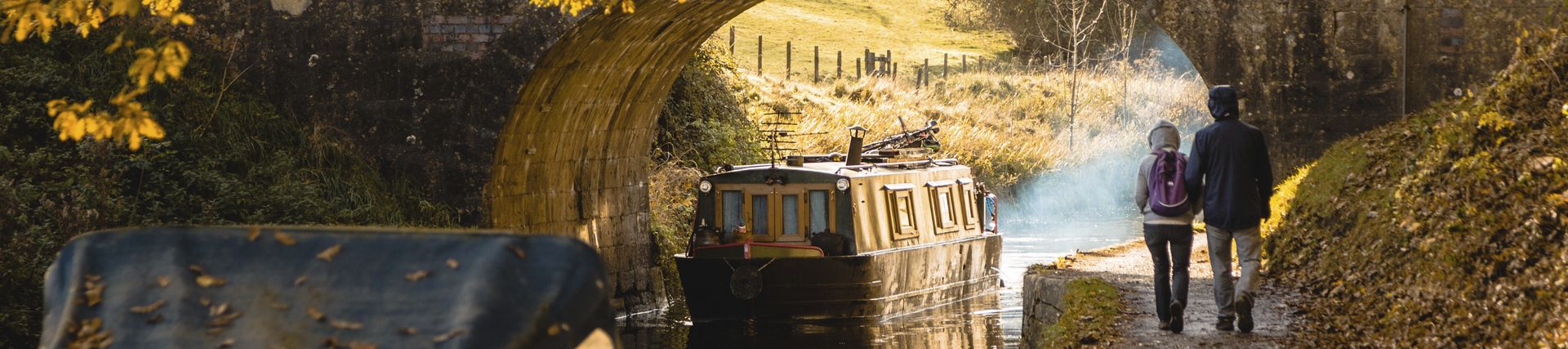 A couple walking beside the canal approaching a low bridge in Bathamption, Somerset, England