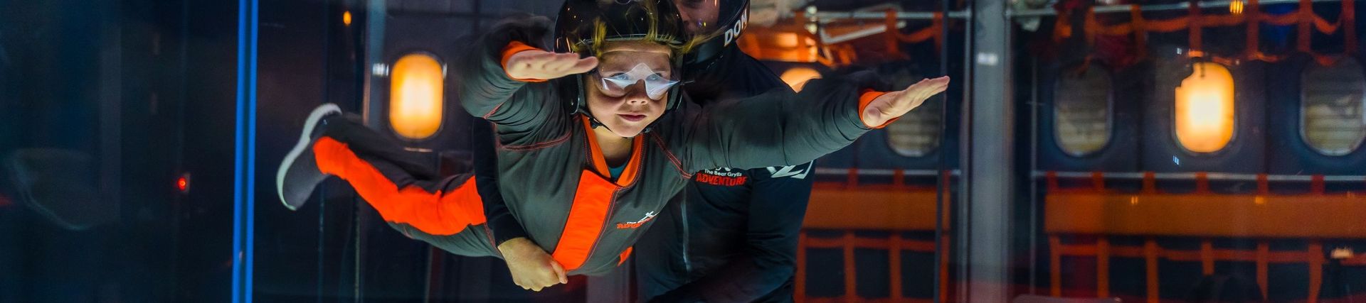 Child in helmet hovering horizontally in the air in a simulator being assisted by an instructor