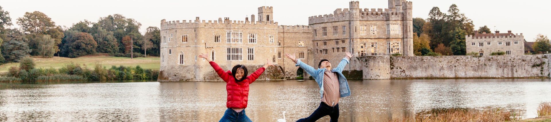 Couple, wearing red jacket, jumping on a lawn by the moat.