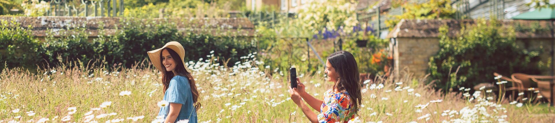 Two women enjoy gardens full of daisies in front of a stone house