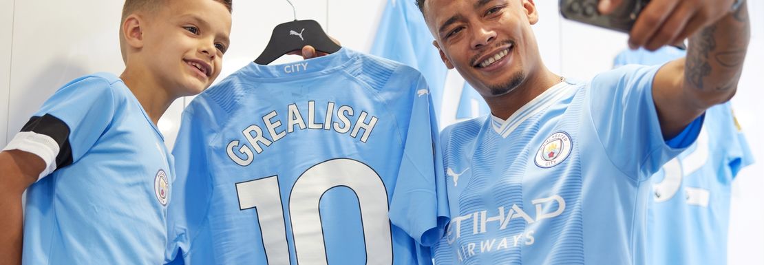 Two Manchester City fans in light blue getting a selfie with a Jack Grealish number 10 shirt