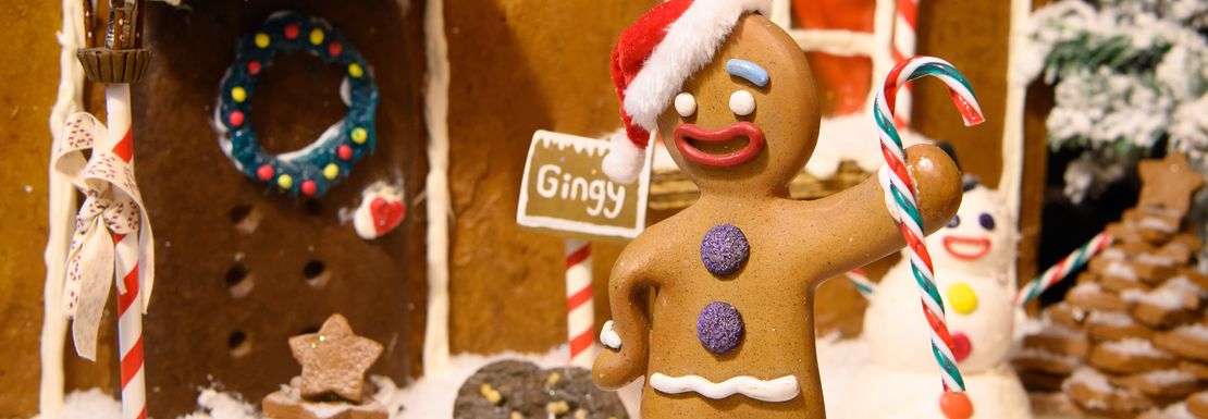 Gingy the Gingerbread Man waves in front of his gingerbread house.