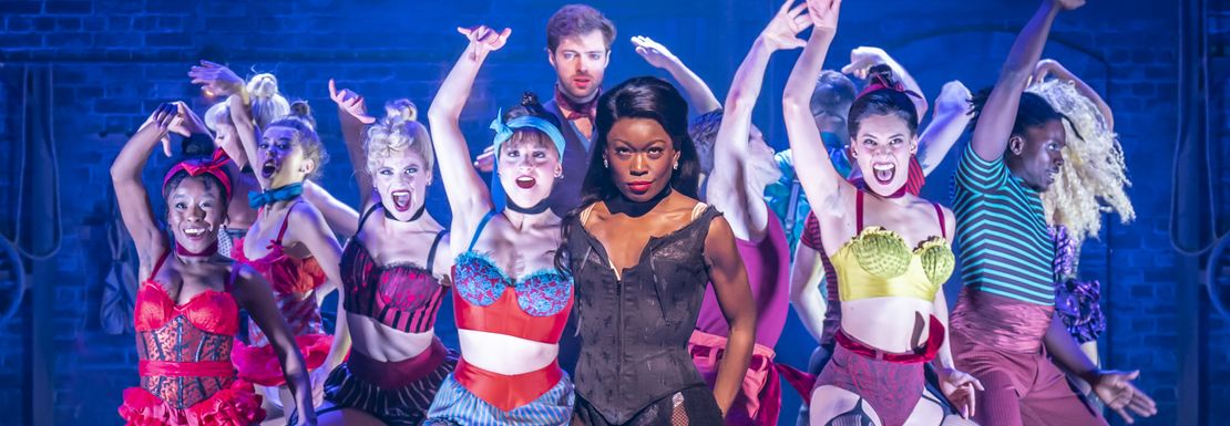 A tow of flamboyant dancers on stage at Moulin Rouge lift their hands in dance
