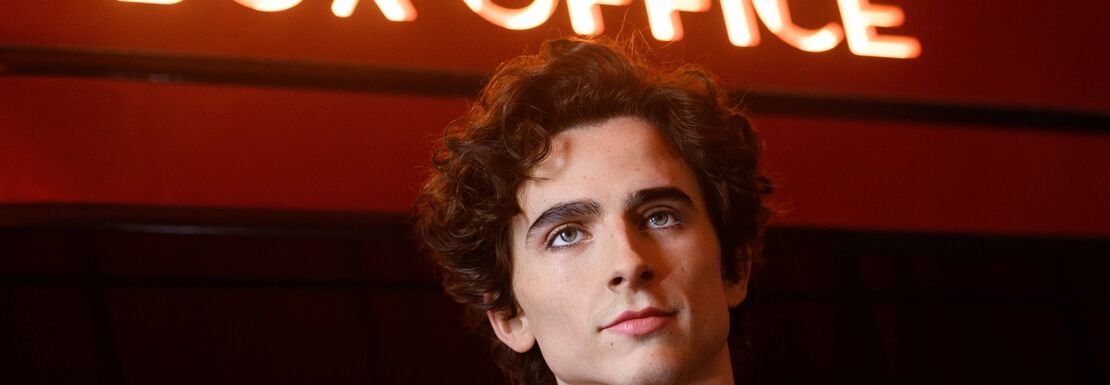 A well-dressed Timothée Chalamet standing underneath a box office sign