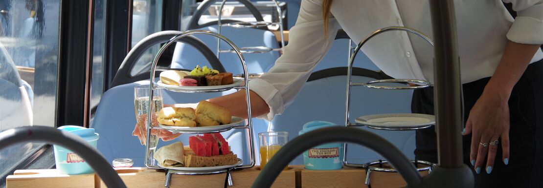 A woman wearing a white shirt set cakes and well presented snacks onto a table on top of a double decker bus.