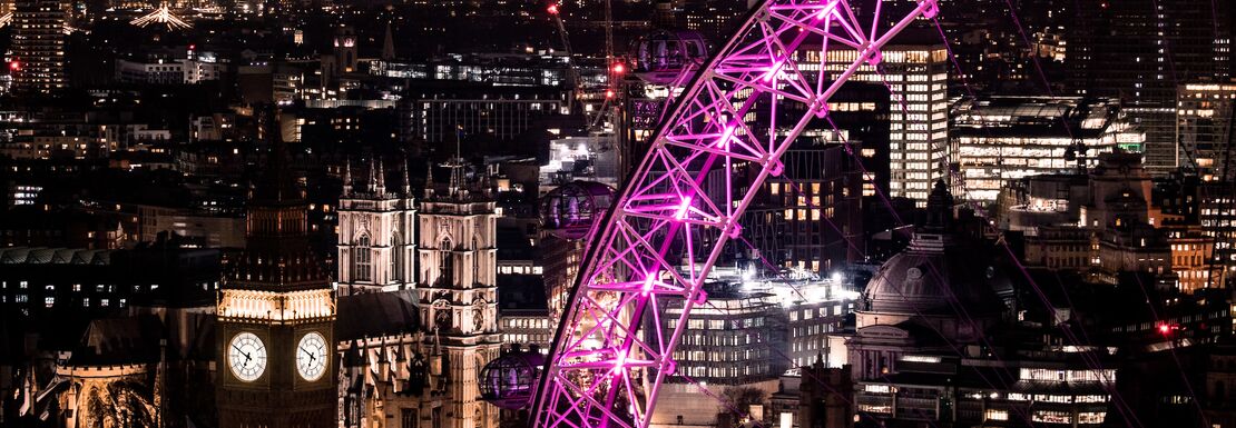 London Eye lit up purple at night over the dark London sky with Big Ben to the side