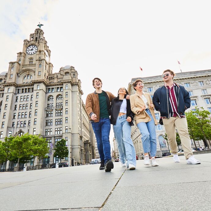 Four friends exploring Liverpool walking along waterfront promenade, with historic buildings in the background